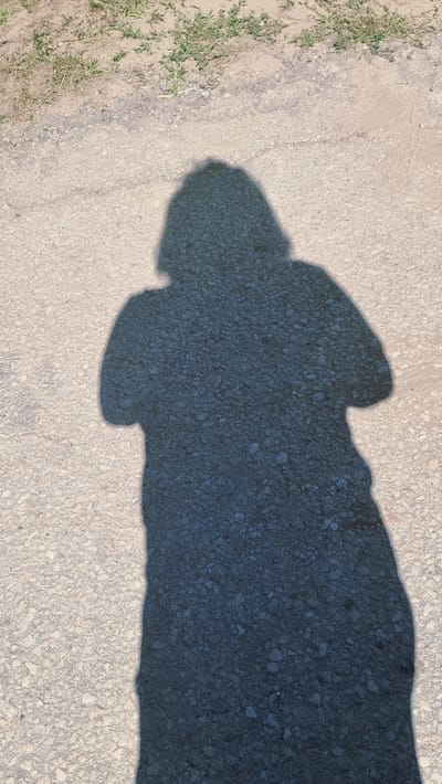A shadow of a person stands on a dirt road in Montagut i Oix, Spain. The shadow is cast from a person with medium-length hair. The ground is composed of light-colored gravel with scattered patches of dry grass in the upper portion of the image. The shadow occupies the majority of the frame, extending from the top left to the bottom right.