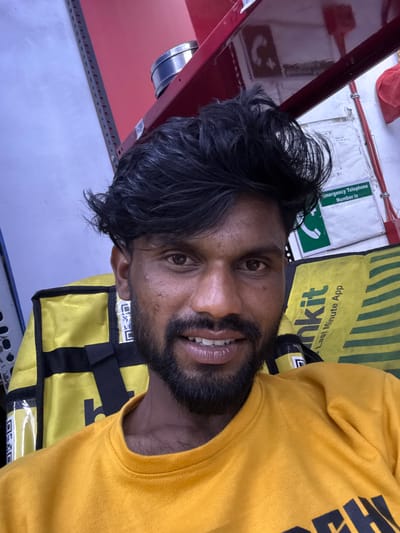 A South Asian male with dark complexion, a full beard, and voluminous dark hair is depicted in a close-up selfie from the chest up, looking directly at the camera with a slight smile revealing his teeth. He is wearing a plain yellow t-shirt. Behind him, a yellow delivery bag with black straps and the text "blinkit" and "Last Minute App" is visible. The background features a light-colored wall, a red shelf or beam overhead, and a silver cylindrical container. To the upper right, a white sign with a green header reading "Emergency Telephone Number is" and a phone icon is affixed to the wall, alongside red cables. The environment appears to be an indoor commercial or storage space, consistent with a delivery service hub, located in the city of Palghar, India.