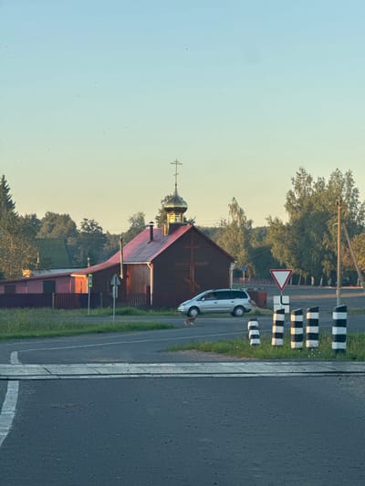 A wide shot captures a rural scene in Ходасаўскі сельскі Савет, Belarus, under a clear, pale blue sky that transitions to a soft yellow near the horizon, suggesting early morning or late evening light. In the mid-ground, a wooden building with a vibrant red roof and a golden dome topped with a cross stands out. This structure appears to be a church or religious building. To the left of the building is a dark red fence.
In front of the building, a silver minivan is parked on the paved road. A small, light-brown dog is visible near the front of the minivan, facing away from the camera. A yield sign, a triangular red-and-white sign, is positioned on a pole to the right of the minivan, near the road. Further to the right, a series of striped black and white bollards are lined up along the edge of the road. The road itself features a pedestrian crossing made of metal grating.
The background is filled with dense green trees, characteristic of a wooded rural landscape. The overall impression is one of tranquility and a typical village setting in the Belarusian countryside.