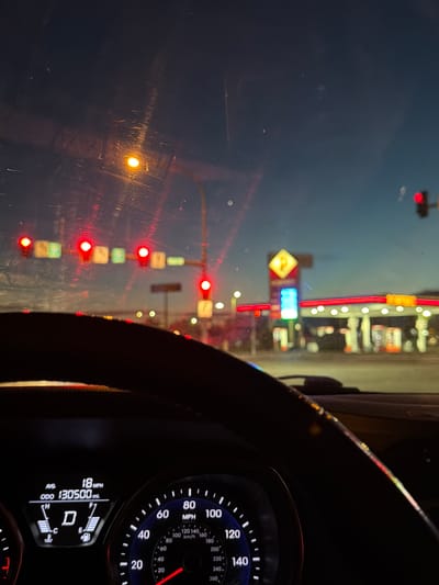 The image is a first-person view from the driver's seat of a car at dusk. The dashboard displays a speed of 18 MPH and an odometer reading of 130,500 miles. Traffic lights are visible ahead, with several showing red. In the background, a gas station with illuminated signage and pumps is present. The overall scene depicts a car driving through a city intersection in Pasco, USA, during twilight hours.
