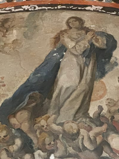 This media file captures a detail from an aged religious fresco or mural, likely dating from the Colonial era, in Tepotzotlán, Mexico. The artwork features a muted color palette dominated by earthy tones, ochres, browns, and deep blues, exhibiting visible signs of wear and cracking consistent with its age.
The central focus is a light-skinned woman with dark hair, dressed in a light-colored garment, depicted in a devotional pose. Her hands are clasped in prayer, and her upward gaze conveys an expression of serenity or spiritual contemplation, suggesting a moment of grace, ascent, or heavenly vision. Behind and partially above her is another figure, heavily draped in a deep blue mantle, a color often associated with the Virgin Mary in Christian iconography. This figure's hands are also joined in a prayerful or supplicatory gesture, though their face is largely obscured.
Surrounding these central figures, particularly on the right and lower right, are numerous cherubic figures (putti). These plump, childlike forms, characteristic of religious art, appear to be floating amidst clouds, reinforcing the ethereal and celestial setting. A faint, warm orange-yellow glow or halo is visible behind the upper portion of the composition, indicating a divine light source. The diagonal arrangement of the figures creates a sense of upward movement or elevation. The scene's iconography strongly suggests a religious narrative such as the Assumption of the Virgin Mary or the glorification of a female saint. No text is visible in the image.