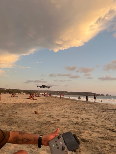 The image depicts a beach scene in Brisas de Zicatela, Mexico, during the late afternoon or early evening. The sky is partly cloudy with warm-toned clouds on the left side and lighter clouds scattered across the center.
A drone is in flight, hovering over a crowded section of the beach. People are sunbathing, walking, and engaging in various activities along the shoreline. Coconuts are scattered on the sand.
In the foreground, a person holds a drone controller attached to a smartphone. The person's arm is visible, adorned with tattoos. The sand is textured and dry near the person, transitioning to a smoother, potentially wetter surface closer to the water. Waves are visible in the background.