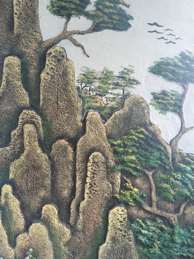 This media file showcases a vibrant, textured relief sculpture or mural depicting a stylized mountainous landscape. The art features prominent, rugged rock formations that dominate the scene, rendered in earthy tones of brown, gold, and dark green, giving them a mossy, aged appearance. The textured surface of the relief impressively mimics the rough, craggy nature of rock and the bark of trees.
Interspersed among the tall, vertical rock peaks are numerous trees. These trees, some resembling pines with their distinct branch structures, are painted in various shades of green, from dark forest green to lighter, almost yellowish-green for the foliage, suggesting highlights or different species. The branches themselves are sculpted in relief, adding to the three-dimensional quality of the artwork.
The upper section of the artwork depicts a clear, bright sky, painted in a light, almost white hue. A small flock of dark, silhouetted birds is visible flying across this expansive sky in the upper right portion of the image, adding a sense of scale and movement to the otherwise static natural scene.
The overall artistic style strongly evokes traditional East Asian landscape paintings, particularly 'shan shui' (mountain-water) art, characterized by its emphasis on mountains, trees, and ethereal compositions. This detailed and tactile artwork is located in Bangsri, Indonesia. There is no visible text in the image. The time of day appears to be bright daytime, under clear skies.