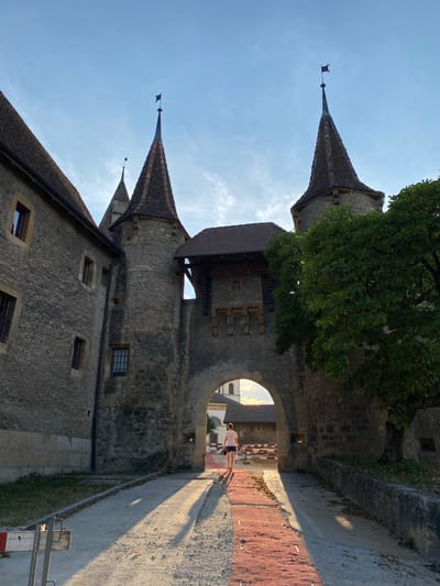 The image shows a person walking through a stone archway with two conical towers on either side, located in Milvignes, Switzerland. The architecture appears to be historic, resembling a castle or fortified building. The person, wearing a white t-shirt and blue shorts, is walking away from the camera towards a building visible through the archway. The sun is casting long shadows across a red brick pathway leading into the arch. Trees and a stone wall are visible to the right of the archway, and part of a stone building with windows is on the left. The sky is blue with some wispy clouds. In the background, there is construction equipment and fencing.