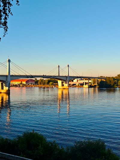 The image shows a view of the Kimry Bridge in Kimry, Russia, as seen from a body of water during sunset. The sky is a clear, gradient blue, transitioning to a pale yellow near the horizon. A suspension bridge with two tall, white pylons dominates the middle ground, with cables stretching to support the roadway. The bridge is illuminated by the warm glow of the setting sun, which casts golden reflections on the rippling water below.
On the far side of the river, behind the bridge, are several buildings, including a large, orange, warehouse-like structure with "RYBPROMTEKHNIKA" written on it. There are also trees and smaller buildings visible. The foreground features dark green foliage, possibly bushes or trees, along a concrete embankment. The water itself is a deep blue, reflecting the sky and the bridge in shimmering patterns. A few small boats are docked near the shore on the other side of the river, and a couple of figures can be seen walking on the bridge. The overall impression is one of a peaceful, late afternoon or early evening scene in a Russian city.