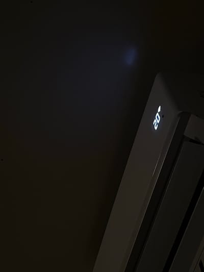 The image shows a close-up of a white air conditioning unit with a digital display showing "20°". The unit is angled, with the display facing upwards and to the left. The background is dark, with a faint blue light emanating from the top left of the frame, possibly from the ceiling. The scene appears to be indoors, in a dimly lit environment. There are no people or specific identifiable landmarks of Genemuiden, Netherlands visible in the image.