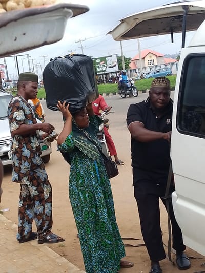 The image depicts a street scene in Oja Oba, Oko Erin, Nigeria. In the foreground, a woman balances a large black plastic bag on her head, dressed in a green and blue patterned garment and a headscarf. To her left, a man wearing a green cap and a patterned brown, blue, and white outfit holds out a wad of Nigerian Naira notes. Behind them, a white van is partially visible, with its door ajar. Another man in a black polo shirt with a white collar and "33 Lacoste" written on it, wearing a patterned cap, stands near the van's door, seemingly interacting with the people. In the background, a motorcycle with two riders passes by. Several streetlights and a billboard with a portrait and text are also visible. The ground is paved with dirt and some concrete. The sky is overcast.