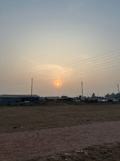 The scene, located in Kumarvog, Bangladesh, depicts an outdoor landscape under a hazy sky during either sunrise or sunset. A distinct orange sun is positioned slightly left of the image center, casting an orange-yellow glow that transitions into a pale blue upper sky. Numerous overhead power lines are visible, supported by multiple dark utility poles stretching horizontally across the mid-upper frame, with small dark objects, consistent with birds, perched on several wires. The foreground consists of dry, brown earth with sparse vegetation, including a section of reddish-brown brick debris or paving in the lower right. Beyond an expanse of barren ground, the background features a horizontal arrangement of diverse low-rise structures; some appear industrial with corrugated metal or tarpaulin coverings, while others resemble makeshift shelters or stalls. To the far right in the background, several vehicles are parked. No individuals or specific actions are discernible.