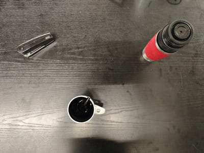 The image displays a top-down view of a dark wooden desk with three objects placed on it: a stapler, a thermos, and a mug of coffee. The stapler, a silver Novus brand, is positioned on the left side of the frame. To its right and further up is a red and black thermos. In the lower left quadrant of the image, a white mug filled with dark liquid, presumably coffee, sits with a spoon resting inside it. The scene appears to be indoors, with the lighting suggesting it could be daytime. There are no people visible. The overall impression is one of a workspace or a casual setting.