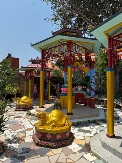 The image shows a street-side view of a Buddhist temple in Unknown, Vietnam. The temple is characterized by traditional Asian architectural elements, including red and yellow support beams, and a multi-tiered roof. The roof is painted with light green and yellow trim. Colorful Buddhist prayer flags are strung along the roofline and between pillars.

Several golden turtle statues are positioned along a walkway made of irregularly shaped mosaic tiles. These statues stand on pedestals adorned with floral designs in pink and green. The pillars leading to the temple are painted gold with red and blue accents.

The temple features intricately carved and painted decorations along the walkway. In the background, there is a glimpse of surrounding buildings and foliage, suggesting an urban setting. The sky is clear blue.