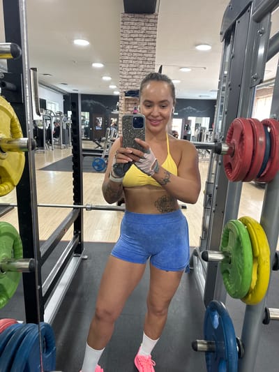 The image shows a woman in a gym, taking a selfie in a mirror. She is wearing a yellow sports bra and blue shorts. She has tattoos on her arms and a ring on her finger. She is also wearing fitness gloves and white socks with pink sneakers. In the background, there are weight plates on a rack, gym equipment, and mirrors. The gym appears to be well-lit with overhead lights. The woman is smiling and looking at the camera. The image is oriented vertically. There is no visible text in the image. The location is Yuzhnouralsk, Russia.