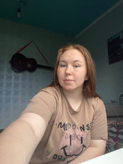A young woman with reddish-brown hair and freckles is taking a selfie. She is wearing a tan t-shirt with black and pink text that reads "MEDOAN SPORT FORCE" and a smiley face graphic. Her gaze is directed towards the camera. The setting appears to be indoors, with a wall covered in a patterned wallpaper. A dark-colored guitar is hanging on the wall to her left, suspended by a red string. A lightbulb is visible in the upper left corner of the frame. The overall lighting suggests it is daytime. The image is angled, as if the woman is holding the camera at arm's length from a low angle.