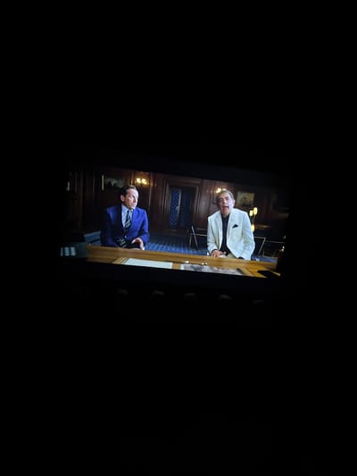 Two men are seated at a wooden desk in what appears to be a formal room with wood-paneled walls. The man on the left wears a blue suit with a green and white striped tie. The man on the right wears a white suit jacket over a dark shirt. Both men are looking towards the viewer. The room has checkered blue carpet and framed pictures on the wall.