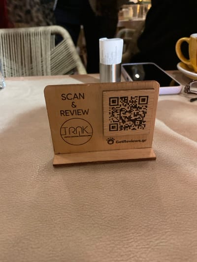 The image presents a close-up, slightly overhead view of a wooden stand placed on a light-colored, textured table, likely in a cafe or restaurant in Chaidari, Greece. The primary subject is this wooden stand, which serves as a customer engagement tool.
Prominently displayed on the stand is a dark QR code. Below the QR code, the phrase "SCAN & REVIEW" is printed vertically. Further down, a circular logo reads "TRNK," with smaller text below it listing "COFFEE | COCKTAIL | BRUNCH | FOOD," indicating the services offered by the establishment. To the right of the QR code, the text "GetReviews.gr" is visible, accompanied by an icon depicting four stars.
Behind the wooden stand, a metallic holder secures a small, rolled-up piece of paper, which appears to be a receipt or bill. Partial Greek text is visible on this paper, including words such as "ΑΠΟΔΕΙΞΗ" (receipt). In the upper left corner, part of a yellow or mustard-colored cup is visible. A smartphone with a reflective screen is positioned behind the receipt holder. The background features blurred elements, including what appears to be woven or cane-back chairs, consistent with an indoor dining or lounge environment. The scene depicts a setup designed to solicit customer reviews digitally.