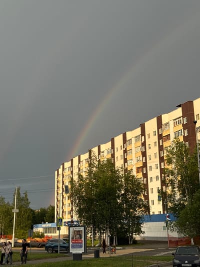 A faint rainbow arcs across a gray, overcast sky above a modern apartment building in Surgut, Russia. The building is cream-colored with dark brown vertical accents and features numerous windows. In the foreground, green trees line the street, partially obscuring the building and the sky. A blue and white storefront, labeled "СПОРТМАСТЕР," is visible behind the trees. Several people are walking on a sidewalk near the building, and a blue car is parked on the street. A billboard displays a couple and Russian text. The scene appears to be an urban residential area during daylight hours.
