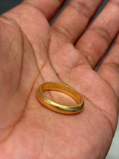 A gold Cartier ring sits in the palm of a brown-skinned hand. The ring’s inner band is engraved with "© Cartier 59 WGW609 A". The hand's lines and texture are visible. The background is a blurry, dark gray. The lighting is diffused, emphasizing the ring's color and the details of the hand.