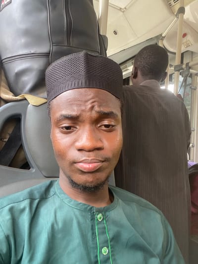 Inside a vehicle, likely a public transit bus in Epe, Nigeria, a young man of dark complexion is depicted in a medium close-up, facing the camera directly. He wears a dark green traditional long-sleeved top with a band collar and light green buttons/trim, along with a black, textured cap covering his head. His expression is neutral with slightly furrowed brows. Behind him, a grey Fila-branded backpack rests on a seat. To his right and slightly behind, another individual with short dark hair is partially visible from the rear, wearing a dark brown garment and holding onto a white overhead grab bar. The vehicle's interior features a white ceiling with ventilation grilles, white vertical and horizontal grab bars, and grey upholstered seats.