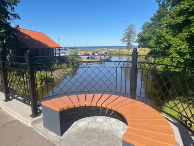 The image depicts a picturesque waterfront scene in Toila, Estonia, on a bright, sunny day. In the foreground, a modern, curved wooden bench with dark grey legs sits on a paved area, offering a vantage point overlooking a calm body of water. A black wrought-iron fence with decorative swirls separates the bench area from the water.
Across the water, a rustic wooden building with a vibrant orange tiled roof is visible, possibly a boathouse or cafe. The water itself is a dark, reflective surface, mirroring the blue sky and the surrounding greenery. A variety of boats, including sailboats with tall masts and smaller motorboats, are moored in the distance, suggesting a marina or harbor.
The background is filled with lush green trees lining the far bank of the water, and the distant horizon shows the expanse of the sea under a clear blue sky. The overall atmosphere is tranquil and inviting, characteristic of a coastal resort town. There are no people visible in the image.