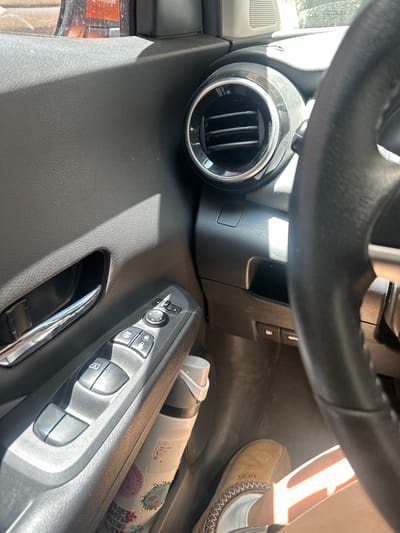 The image provides an interior view of a car, observed from the driver's perspective, looking towards the passenger side and down. The scene is illuminated by bright, direct sunlight streaming into the vehicle, creating strong highlights and distinct shadows, suggesting a sunny daytime in Jesús del Monte, Mexico.
A section of a black, textured steering wheel occupies the top of the frame. Below it, on the dashboard to the left, is a circular air vent with a metallic trim and vertical slats. The dashboard and door panels are made of black, textured plastic.
Prominently featured on the door panel is a control module for various functions. It includes buttons for power windows, one marked with an "A," suggesting an automatic function, and others for door locks, indicated by lock and unlock symbols. A joystick-like control for mirror adjustments is also visible. Below these controls, a gleaming silver door handle is partially seen.
On the passenger side floor, a person's foot is visible, clad in a light brown, moccasin-style slipper. The "UGG" logo can be faintly discerned on the side of the shoe. Adjacent to the shoe, a white bottle or tumbler, decorated with several colorful stickers, lies on the floor. Visible text on these stickers includes "GROOVY" and part of another phrase appearing as "JOY WITH," accompanied by various floral and abstract designs.
No particular activity or event is depicted, other than the car being occupied. The overall impression is a casual, everyday moment inside a vehicle.