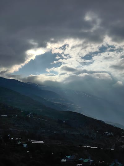 This is a vertical, eye-level, medium shot of a mountain landscape in the vicinity of Gondhla, India. The upper half of the image is dominated by a dramatic sky with dark, heavy clouds partially obscuring a bright sun. Streaks of sunlight break through the clouds, illuminating parts of the sky with a soft glow. Below the sky, the dark, steep slopes of mountains recede into the distance. The mountains are covered in vegetation, with some patches of snow visible on the higher peaks. Scattered across the lower slopes are small clusters of buildings, some appearing to be houses with green roofs, and several long, white greenhouse-like structures. The overall mood is somber and atmospheric, with a sense of isolation and rugged natural beauty.
