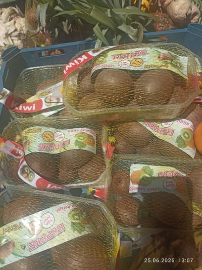 The image shows a close-up, overhead view of several packages of kiwi fruit inside a blue crate. The kiwi are encased in transparent plastic packaging, with a gold mesh net covering them. Each package has a colorful label featuring text in Russian, including "БЕРЕКЕ" (BEREKE), "Свежие киви" (Fresh kiwi), and "FOR EXPORT". There are also illustrations of kiwi fruit on the labels. In the background, there are glimpses of other fruits and greenery, possibly pineapples, and a portion of a brown, textured surface that could be a table or counter. The scene appears to be in an outdoor market or grocery setting, with natural light illuminating the produce. The timestamp "25.06.2026 13:07" is visible in the lower right corner of the image. The image does not provide specific details about the location within Razzakov, Kyrgyzstan, beyond it being a place where kiwis are sold.