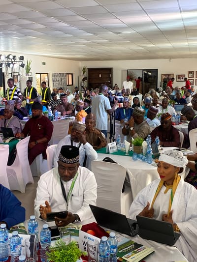 A photograph depicts a meeting or conference taking place indoors in what appears to be Kwarau, Nigeria. The room is populated by many people, mostly men, seated at tables covered with white tablecloths. The majority of the attendees are wearing traditional Nigerian attire, including hats, robes, and gowns. Several individuals have laptops and water bottles in front of them, and a few are engaged in using their mobile phones or tablets.
The backdrop includes photographs on the wall, indicating a formal setting. In the foreground, there are multiple water bottles placed on the table. The lighting in the room is bright, suggesting an indoor setting. The individuals present seem to be participating in a professional event, likely related to agricultural matters indicated by documents with the words "Mid Term Review," and "World Bank."
