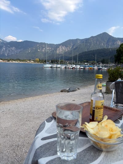This is a medium-shot, sideways photograph taken outdoors. The scene features a body of water, likely a lake, with a mountain range in the background. Several sailboats are moored on the water. In the foreground, on a gravelly surface, are a clear glass with water, a glass bottle with a yellow cap and label, and a clear glass bowl filled with potato chips. A wooden cutting board is underneath the bottle and bowl. A patterned fabric, possibly a tablecloth or blanket, is laid out on the gravel. The sky is blue with scattered white clouds. The lighting suggests it is daytime, with the sun casting shadows. The setting appears to be a lakeside leisure area. The label on the bottle is partially visible and reads "LURO" and "CHINO". There is no clear indication of people or a specific event taking place, suggesting a moment of relaxation.