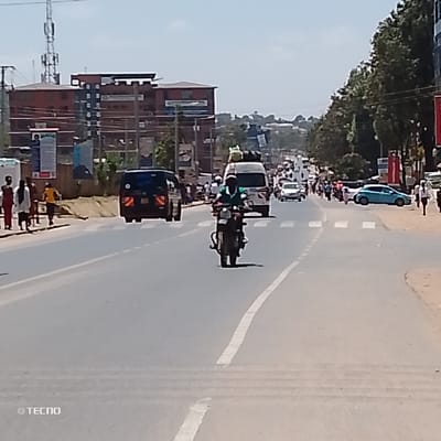 The image depicts a daytime street scene in Machakos Central ward, Kenya, looking down a multi-lane asphalt road that recedes into the distance. The sky is clear, indicating sunny conditions.
In the immediate mid-ground, a person on a dark motorcycle, wearing a green shirt, is traveling away from the viewer in the center lane. Ahead of the motorcycle and slightly to its right, a white passenger van (likely a matatu) with luggage secured on its roof is also moving away. Behind the motorcycle, a dark-colored SUV or van with rear markings (possibly a police vehicle) is visible, also traveling away.
The road features clear white lane markings, including a solid line on the right side and a broken line indicating a lane boundary or center. A pedestrian crosswalk with white zebra stripes is visible across the road.
On the left side of the road, a dirt shoulder and a raised earth/concrete barrier separate the road from an area with several pedestrians. Multiple individuals are visible here, some walking, including one wearing a yellow shirt. Further back on the left, a prominent multi-story reddish-brown building with numerous windows stands, partially obscured by utility poles and power lines. A tall cellular lattice tower is visible in the far left background.
On the right side of the road, a dirt shoulder borders a tree-lined area. Further down the road on the right, a light blue sedan is parked or stationary. Numerous pedestrians are also visible on the right side of the road in the distance, and several other vehicles are in various stages of traffic, extending towards the horizon where the road appears to ascend. Commercial signage and buildings are partially visible behind the trees on the right.