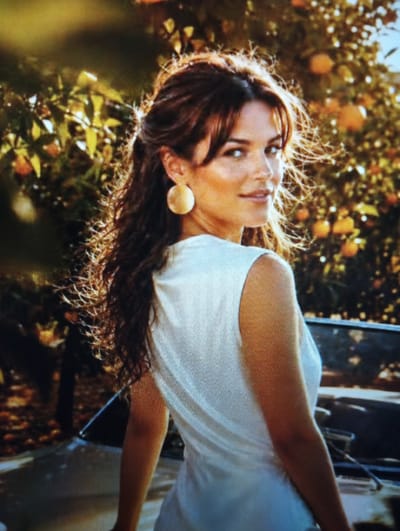 The image shows a woman with long, dark, wavy hair, wearing a white sleeveless top and a large, round, gold earring. She is looking over her left shoulder towards the camera with her head tilted slightly. Her hair is blowing in the wind. She is outdoors, with a background of trees with oranges or similar fruit, and dappled sunlight. A convertible car is partially visible in the foreground. The image does not provide any information about the city of Kingisepp, Russia.