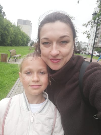 A woman and a young girl are taking a selfie in an outdoor setting. The woman is on the right, and the girl is on the left, slightly in front of her. They are both looking at the camera. The woman has dark hair pulled back, and is wearing a dark brown turtleneck sweater. The girl has blonde hair and is wearing a white quilted jacket over a white shirt. She has glitter applied to her cheeks. In the background, there is a park-like area with green grass, trees, a wooden bench, and a trash can. Behind the park, there are residential buildings. The sky appears overcast, suggesting it might be a cloudy or early evening. The overall mood seems casual and familial. There is no visible text in the image.