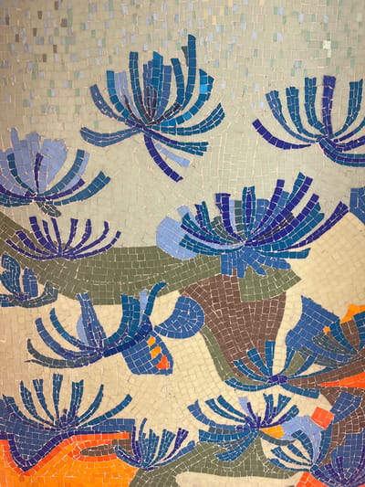 This is a close-up, overhead photograph of a mosaic artwork. The mosaic is composed of small, square tesserae in shades of blue, green, brown, orange, and cream. The artwork depicts abstract floral or botanical forms, characterized by segmented blue petals radiating outwards from a central point. Some of the forms are interspersed with areas of solid green and brown tesserae, possibly representing leaves or earthy elements. Bright orange tesserae appear in scattered areas, adding pops of color. The background is a uniform cream color with subtle variations in tone. The mosaic appears to be a wall or floor covering.

The mosaic is located on the exterior wall of the L'Espace Decoratif of the Madoura pottery complex in Vallauris, France. This complex was famously associated with Pablo Picasso and his ceramic work.