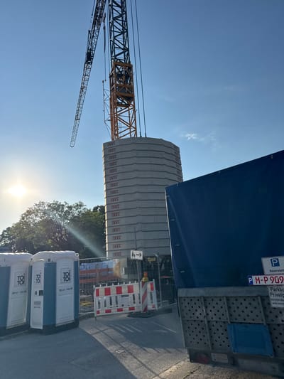 This image captures a construction site bathed in bright sunlight, likely during the late morning or early afternoon. The clear blue sky dominates the upper portion of the frame, with only a few wispy clouds visible.

The foreground features several portable toilets, branded with "TOI TOI". One of these is identified with the number "08141-536 80".

Further back, a large, imposing stack of concrete barriers, each marked with "BKL", forms a significant part of the scene. These barriers are positioned next to a construction crane, which is partially visible against the sky. The crane's yellow and grey structure suggests active construction work.

To the right, a blue tarp covers a portion of a trailer or vehicle. A sign with a parking symbol and the text "P Reserviert" (Reserved) is visible on the trailer, along with "PM-P 909" and "Parkbuch Privato" (Private parking). Additional text below indicates "Widerrechtlich abgestellte Fahrzeuge werden kostenpfl...".

A road or paved area is visible to the right of the portable toilets, with shadows cast across it, suggesting the sun's position. The presence of construction equipment and barriers strongly indicates an ongoing construction or roadworks project. The overall atmosphere is one of industry and development under a clear, sunny sky.