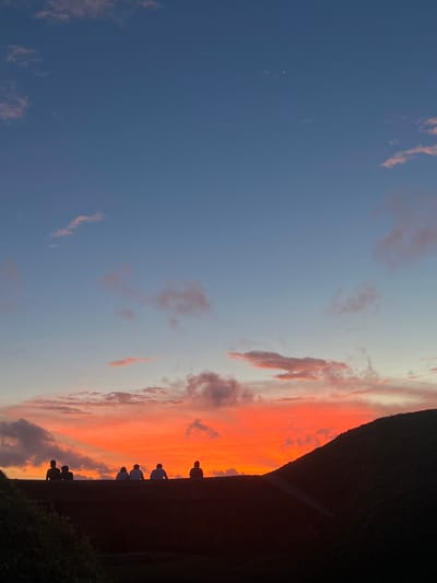 The image depicts a group of silhouetted figures standing on a wall, watching a vibrant sunset over Galle, Sri Lanka. The sky is ablaze with fiery orange and pink hues, a classic sunset palette that signals the end of the day. The clouds are scattered, some tinged with the sunset's colors, while others are darker and more defined. The figures, seen from behind, are gathered together, their forms stark against the bright sky, suggesting a shared experience of observing the natural spectacle. The dark, textured wall they are perched on provides a strong horizontal line, contrasting with the expansive sky. The presence of foliage at the bottom right corner hints at an outdoor setting, possibly a viewpoint overlooking the ocean or a cityscape. The overall mood is one of peaceful contemplation and appreciation for the beauty of nature. The time of day is clearly dusk, with the sun having recently set. There is no visible text in the image.