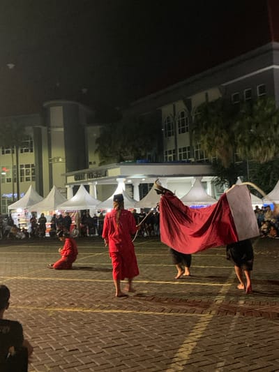 The image shows a performance in an outdoor space in Kecamatan Batu, Indonesia, at night. There are a group of people, some in red costumes and others in black, performing a traditional dance. In the foreground, two men are holding a large red cloth, possibly a costume, as a third man kneels on the ground. The performance is taking place in front of a large building with windows and a white awning. The building appears to be a government building. There are numerous white tents in the background, and the space is illuminated by streetlights. The ground is paved with red bricks.