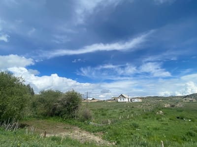 The image captures a rural landscape in Kara-Oi, Kyrgyzstan, under a vast, dynamic sky filled with a mix of white cumulus and wispy cirrus clouds. In the foreground, lush green grass covers rolling hills, interspersed with a few small wooden fences. To the left, dense green bushes and trees dominate, with a small cultivated patch of dark soil visible behind a row of thin, upright sticks.
Further into the scene, several simple houses with white walls and dark roofs are scattered across the landscape. One prominent house, with a pitched roof and visible windows, stands out against the green terrain. Beyond the houses, the land continues to roll, with some areas appearing dry and barren. A few more structures and fences are visible in the mid-ground and background, suggesting a sparsely populated rural settlement. The overall impression is one of a quiet, open countryside typical of a village environment in Kyrgyzstan.