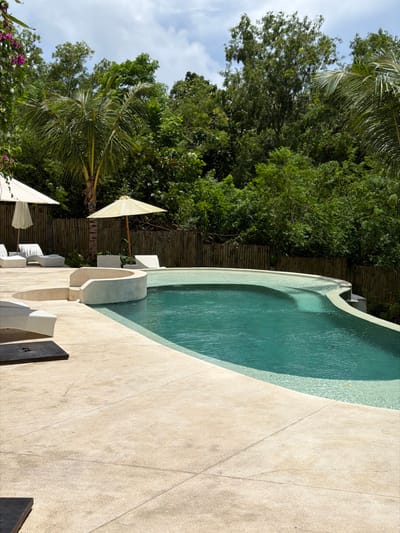 A curved swimming pool with clear water sits next to a light beige concrete patio. Lounge chairs with white cushions are arranged near the pool. Behind the patio, a bamboo fence separates the area from a lush green forest with palm trees. White and beige umbrellas provide shade. The sky is visible with scattered clouds.