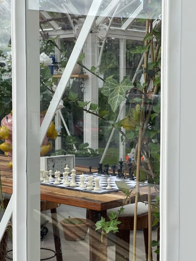 The image shows a view through a glass window of a greenhouse located at 64.139 latitude and -21.931 longitude in Reykjavík, Iceland. Ivy plants grow outside the greenhouse and are visible through the glass. A chessboard with white and black pieces set up on a wooden table is inside the greenhouse. A white clock is sitting next to the board, but the time cannot be read from the image. The greenhouse is made of white metal frame with glass panels. The floor of the greenhouse is made of stone. The white window frame is visible in the foreground of the image. Sunlight is shining in the image.