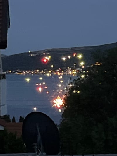 In Baošići, Montenegro, at dusk or during twilight, a scene unfolds with a body of water reflecting lights from a distant shoreline. The water is calm, creating a smooth surface that mirrors the illuminated elements. On the left side of the frame, a dark, satellite dish points towards the sky, positioned in the foreground. Behind it, the edge of a building with a red-tiled roof is visible. The background is dominated by a dark, hilly landscape, possibly mountains, where scattered lights suggest a settlement or buildings. The sky is a deep, muted blue, indicating the transition from day to night. Sporadic bursts of light, resembling fireworks, are scattered across the middle ground, some appearing as vibrant red and yellow orbs, while others are more diffused. These lights are reflected on the water, creating shimmering patterns. Dense, dark foliage occupies the right side of the frame, partially obscuring the view of the distant lights and landscape. The overall impression is one of a serene evening event with a display of lights against a natural and built environment.