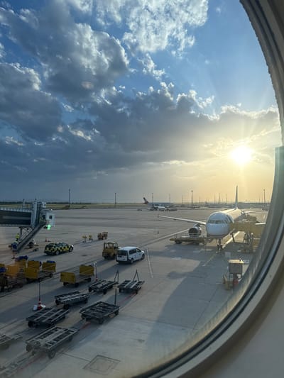 The image shows an airport apron scene viewed through an airplane window. An aircraft is parked at a gate, with ground service equipment and personnel visible around it. The sky is visible with a mix of clouds and sunlight.
Location is likely the Vienna International Airport in Schwechat, Austria.
