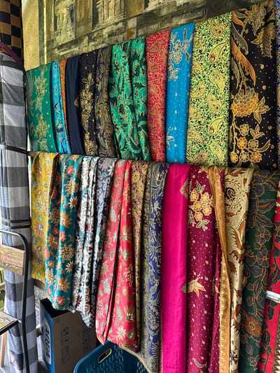 This image captures a vibrant display of batik fabrics hanging on a rack against a textured wall in Tampaksiring, Indonesia. The fabrics are arranged in vertical rows, showcasing a rich variety of colors and intricate patterns. Dominating the scene are the batik cloths, featuring motifs such as flowers, paisley designs, birds, and geometric shapes. The colors range from deep blues, greens, and purples to bright yellows and pinks, often contrasted with gold and black. Some fabrics have a black background with gold motifs, while others are a riot of color with less contrast.
On the left side of the image, a partially visible black and white checkered textile is hung, possibly a sarong or similar garment. Below the hanging fabrics, a blue plastic basket is visible, along with a brown cardboard box that has a prominent blue Aqua logo and some pictograms printed on it. The wall behind the fabrics appears to be made of stone or concrete, with a rough, weathered texture, and a decorative mural or carving of what looks like ancient architecture. The overall impression is of a marketplace or shop stall specializing in traditional Indonesian textiles. There are no people visible in the image.