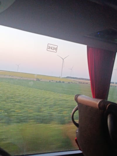 The image is a photograph taken from inside a vehicle, looking out a window at a rural landscape. The time of day appears to be either sunrise or sunset, as the sky is a soft, pale color. In the distance, several wind turbines are visible, dotted across fields of green and yellow crops. The foreground of the image shows the interior of the vehicle, with a seat and a red curtain partially obscuring the view. A sign with the text "16 A16 B" is visible on the window frame. The image is slightly blurred due to motion, suggesting the vehicle is in transit. The location is identified as Poruchik Chunchevo, Bulgaria.