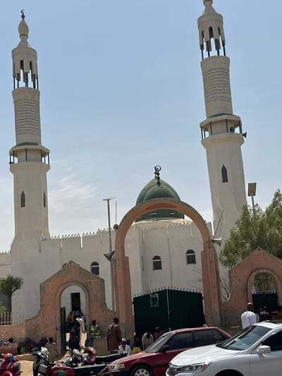The image depicts a bustling street scene in Kofar Ruwa, Nigeria, dominated by a large mosque under a clear, bright sky. The mosque features light-colored, possibly white or cream, plastered walls topped with a crenellated parapet. Two tall, slender minarets stand prominently, one on the left and one on the right, both capped with distinct finials incorporating a crescent moon symbol. The minarets have open upper sections with vertical bars and a lower balcony-like structure fitted with horn loudspeakers. A prominent green dome, also topped with a crescent moon finial, is visible behind a large, decorative archway at the mosque's entrance.
At ground level, the mosque is accessed through multiple arched entryways, some with decorative brickwork patterns in shades of reddish-brown. One such archway in the foreground leads into a courtyard or interior space where numerous individuals are visible, mostly men dressed in traditional Nigerian attire. Many people are gathered around the mosque entrances, with some seated on the dusty ground.
The foreground of the scene is busy with parked and possibly moving vehicles. Multiple motorcycles, including red and green models, are present on the left. A red sedan and a white sedan are partially visible on the right, indicating a busy thoroughfare or parking area in front of the mosque. The ground appears unpaved in some sections. Utility poles with what appear to be streetlights or solar lighting fixtures are visible between the mosque and the foreground. Sparse green trees are also present on both sides of the mosque structure. No specific actions or interactions beyond general gathering and movement are discernible.