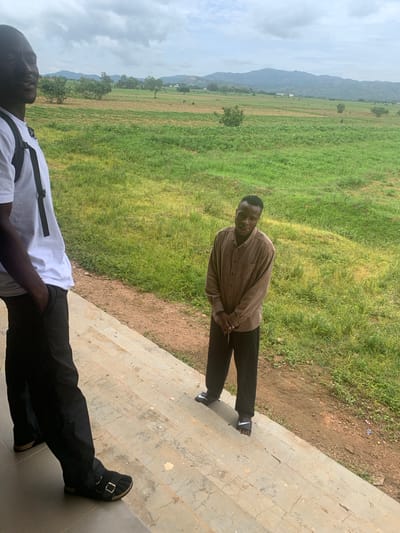 This media file captures two individuals in an outdoor setting. The foreground is dominated by a person wearing a white t-shirt and black trousers, with a backpack visible. Their lower body and feet are primarily in focus, with one foot shod in black sandals. Behind them, a second person stands, wearing a brown checkered long-sleeved shirt and black trousers. They are leaning slightly and looking towards the camera with a relaxed expression.
The background reveals a lush green field stretching towards a range of hills or mountains under a cloudy sky. The presence of scattered trees and what appear to be small structures in the distance suggests a rural or semi-rural environment. The ground near the subjects consists of a concrete staircase or pathway adjacent to the grassy area.
The lighting suggests it could be daytime, with the overcast sky indicating potentially cloudy weather. The overall impression is a casual moment captured outdoors, possibly in a natural or semi-developed area. No visible text is present in the image. The location context provided is Gwafan, Nigeria.