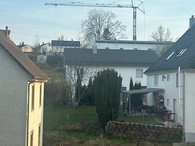 The image presents an overview of a residential area in Krauchenwies, Germany, under an overcast or hazy sky during daylight. The perspective is from an elevated vantage point, looking across a sloped terrain towards distant hillsides.
In the immediate foreground, to the left, a partial view of a light-colored stucco building with a brown-tiled roof and a dark gutter is visible, showing two narrow, vertically oriented windows with brown frames. The central foreground features a sloping grassy area with sparse vegetation and a wire fence. Below the fence, a retaining wall composed of stacked, weathered wood or concrete blocks is present.
The midground is characterized by multiple residential structures. A prominent white, two-story house with a dark gray gabled roof stands centrally, flanked by a tall, dense evergreen shrub on its left. To the right of this house, another white residence with a dark gabled roof displays skylights and a vertical white exhaust pipe on its exterior. Its backyard contains a metal-framed canopy or carport structure, beneath which various items, including stacked materials and what appears to be outdoor equipment, are stored. A third white house with a dark gray gabled roof is positioned further back and to the left of the central white house. Deciduous trees with bare branches are interspersed throughout the scene, particularly between and behind the houses, alongside some evergreen trees.
In the background, additional residential buildings are visible on a higher slope. Dominating the upper central portion of the frame is a large, yellow-and-gray tower crane, its horizontal jib extending across the skyline, indicating ongoing construction activities. No individuals are visible within the depicted scene.
