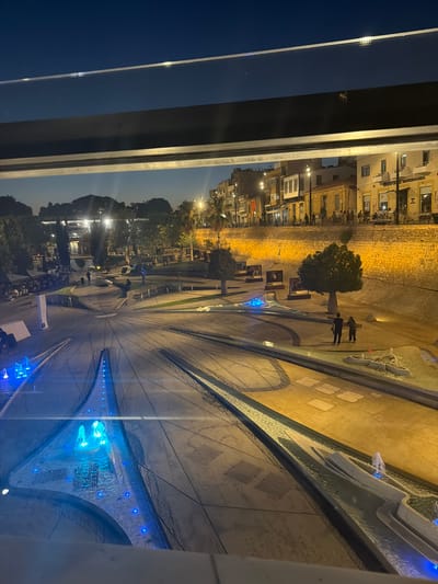 The image depicts a nighttime scene in Nicosia, Cyprus, looking out from a window. The view shows an outdoor public space, possibly a plaza or park, illuminated by artificial lights.

In the foreground, there are water features with blue LED lights, creating a vibrant glow. The architecture visible in the background consists of buildings with warm yellow lighting, suggesting shops or residences. The time of day is clearly night due to the dark sky and the artificial lighting dominating the scene.

There are a few people visible in the distance, walking through the illuminated space. One small group of two or three individuals stands near a tree. Another person appears to be walking further down the path. The overall atmosphere seems calm and serene, with the gentle illumination of the water features and the buildings creating a welcoming ambiance.

The image is taken from a slightly elevated perspective, looking down and across the scene. The framing of the shot suggests the viewer is indoors, looking out through a window, with a dark frame element present on the left side of the image. There is a visible sign on one of the buildings in the background that reads "BIC APOTHEKE". The weather appears to be clear, as there are no visible signs of rain or fog.