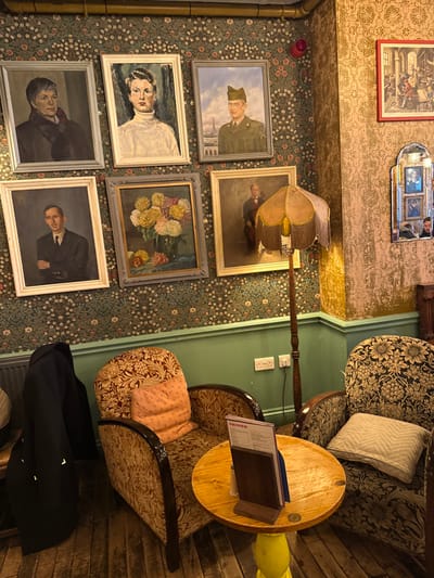 This image depicts an interior section of The Tudor of Lichfield, located in Lichfield, United Kingdom. The scene features a wall adorned with multiple framed artworks and two distinct wallpaper patterns: a dark green floral motif on the left and a gold textured pattern on the right. A green painted dado rail separates the lower wall from the upper.
Six framed paintings are displayed on the floral wallpaper. In the top row, from left to right, are a portrait of a woman with short dark hair, a portrait of a woman in a white turtleneck, and a portrait of a man in a military uniform with the Eiffel Tower in the background. In the bottom row, from left to right, are a portrait of a man in a dark suit with crossed arms, a still life painting of yellow and pink flowers in a vase, and a portrait of a man in formal attire with a musical instrument. A red circular fire alarm device is mounted on the floral wallpaper.
On the right wall, a larger red-framed picture of a historical interior scene is visible above an ornate, arched mirror reflecting a partial human face and additional small framed pictures.
The foreground contains a seating arrangement with two upholstered armchairs flanking a small, round wooden table with a yellow pedestal base. The left armchair is covered in a red and gold floral fabric and holds a pink cushion, with a dark blue/black coat draped over its armrest. The right armchair features a darker floral fabric and a light grey cushion. A wooden menu holder displaying a "DRINKS" menu rests on the table. Between the armchairs, a tall wooden floor lamp with an illuminated, fringed beige fabric shade stands. The floor is composed of dark wooden floorboards. Two white double electrical outlets are visible on the green dado rail. A radiator is partially visible on the far left. No individuals are seated in the immediate foreground.