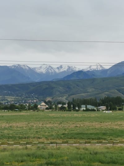 The image depicts a wide landscape under a cloudy sky. In the foreground, a grassy field stretches out, with a low barrier fence composed of alternating dark and light brown and white sections running horizontally across the middle ground. Behind the field, a scattering of buildings, including a large house with a light-colored facade and a green-roofed structure, are nestled amongst trees. Further back, rolling green hills rise, leading to a dramatic mountain range in the distance. The mountain peaks are capped with snow, contrasting with the green slopes below. The overall impression is one of a rural or semi-rural setting in a mountainous region. The sky is overcast, suggesting a potentially cool or dull day. No people are visible in the image. There is no visible text.