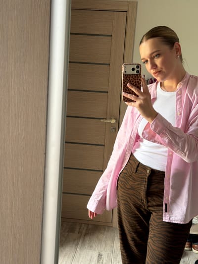 A young woman stands in front of a mirror, taking a selfie. She is wearing a pink button-down shirt over a white tank top and brown zebra-print pants. Her hair is pulled back in a ponytail, and she is wearing subtle makeup. The background shows a wooden door and a wall, suggesting an indoor setting, likely a home. The image does not provide any specific clues or context to pinpoint the location within the city of Dreiliņi, Latvia.