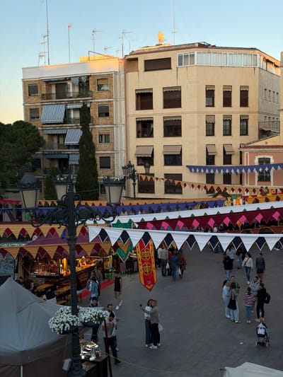 The image depicts a street scene in Burjassot, Spain, evidenced by the buildings and signage. A fair or market is taking place, as indicated by the colorful triangular bunting strung overhead. People are walking through the area, some appearing to be browsing stalls beneath the bunting. The architectural style of the buildings suggests a European urban setting. A lamppost adorned with flowers is visible in the foreground.