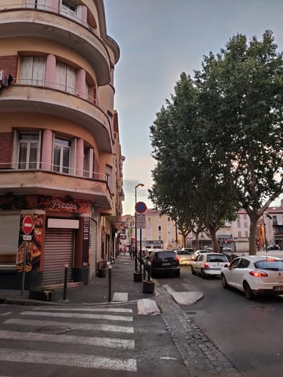 This media file depicts a street scene in Perpignan, France, at dusk. The foreground features a pedestrian crossing with visible zebra stripes and a manhole cover. To the left, a building with rounded balconies and pastel-colored pillars dominates the scene. A storefront displays signage for "Pizzas" and mentions various food items like "kebab," "tacos," and "tajine."
The street is lined with several large trees casting shadows. A line of cars is stopped or moving slowly in traffic, with a white Renault Megane in the right lane being a prominent object. Behind the trees, buildings with windows and awnings suggest a commercial and residential area.
Visible text includes "Pizzas," "kebab," "tacos," "grillades," "tajine," "Bon Appetit," "SAUF MARCHE," "ARMACIE," "Casa Cafe," and license plate details like "CC-161-TF" and "OR-09". The sky is a soft gradient of blue and grey, indicating the time of day is early evening. The overall atmosphere is that of a typical European street during a quiet evening hour, with a mix of commercial activity and residential buildings. There are no discernible people prominently featured. The weather appears calm.